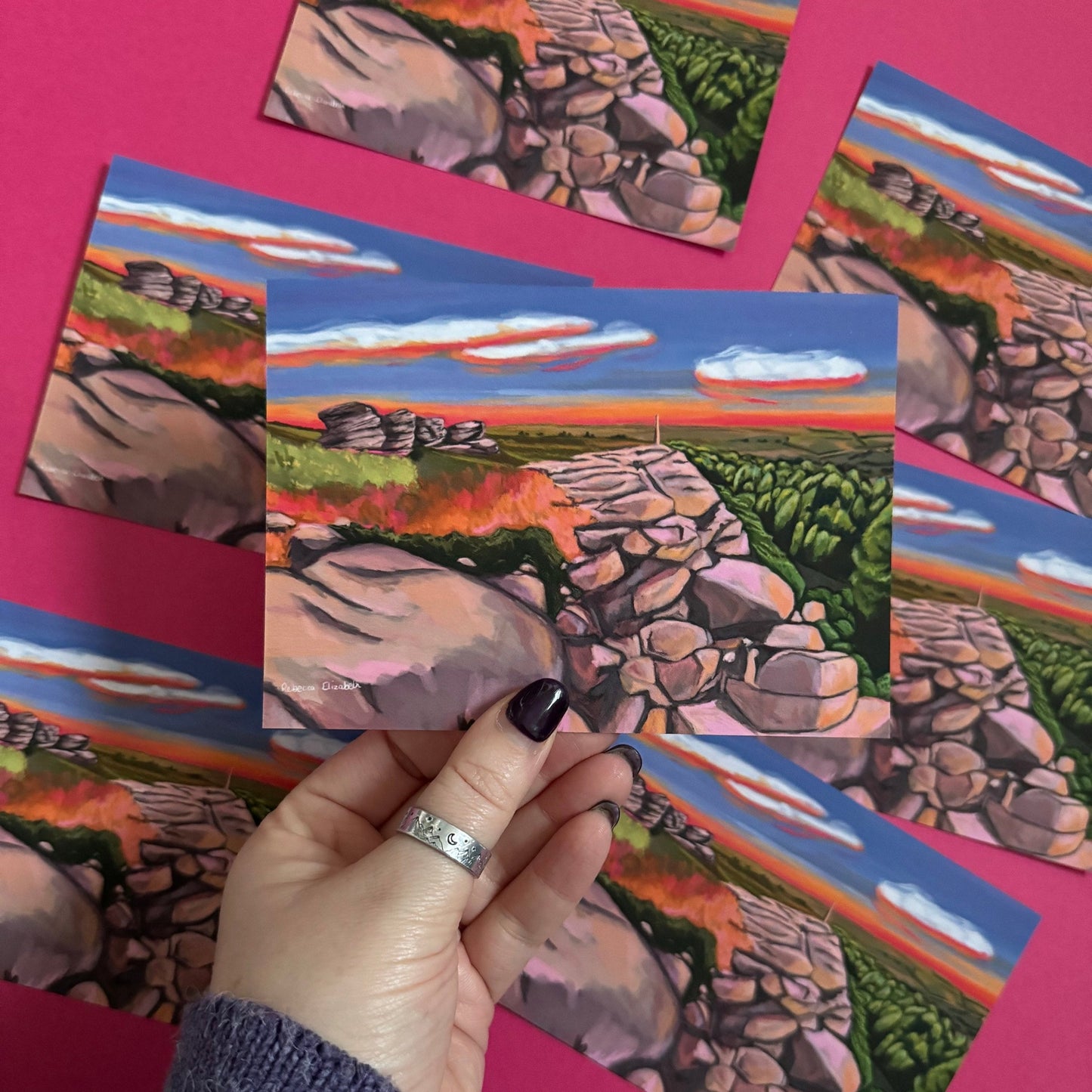 An illustrated postcard of Birchen Edge in the Peak District. With the rock face, rock outcrops named after ships from the battle of Trafalgar and Nelsons Monument all set against a sunset sky with blue, yellow, orange, and pink glow.