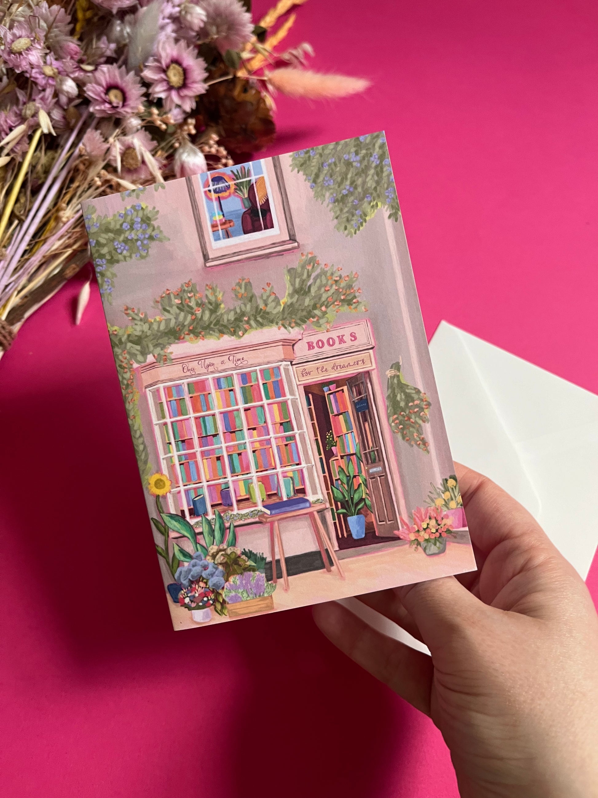 An illustration of a cute whimsical bookshop, the window shows the bookshelves inside full of bright coloured book spines with lots of flowers. The back of the card is pink with the Rebecca Elizabeth Draws logo. 