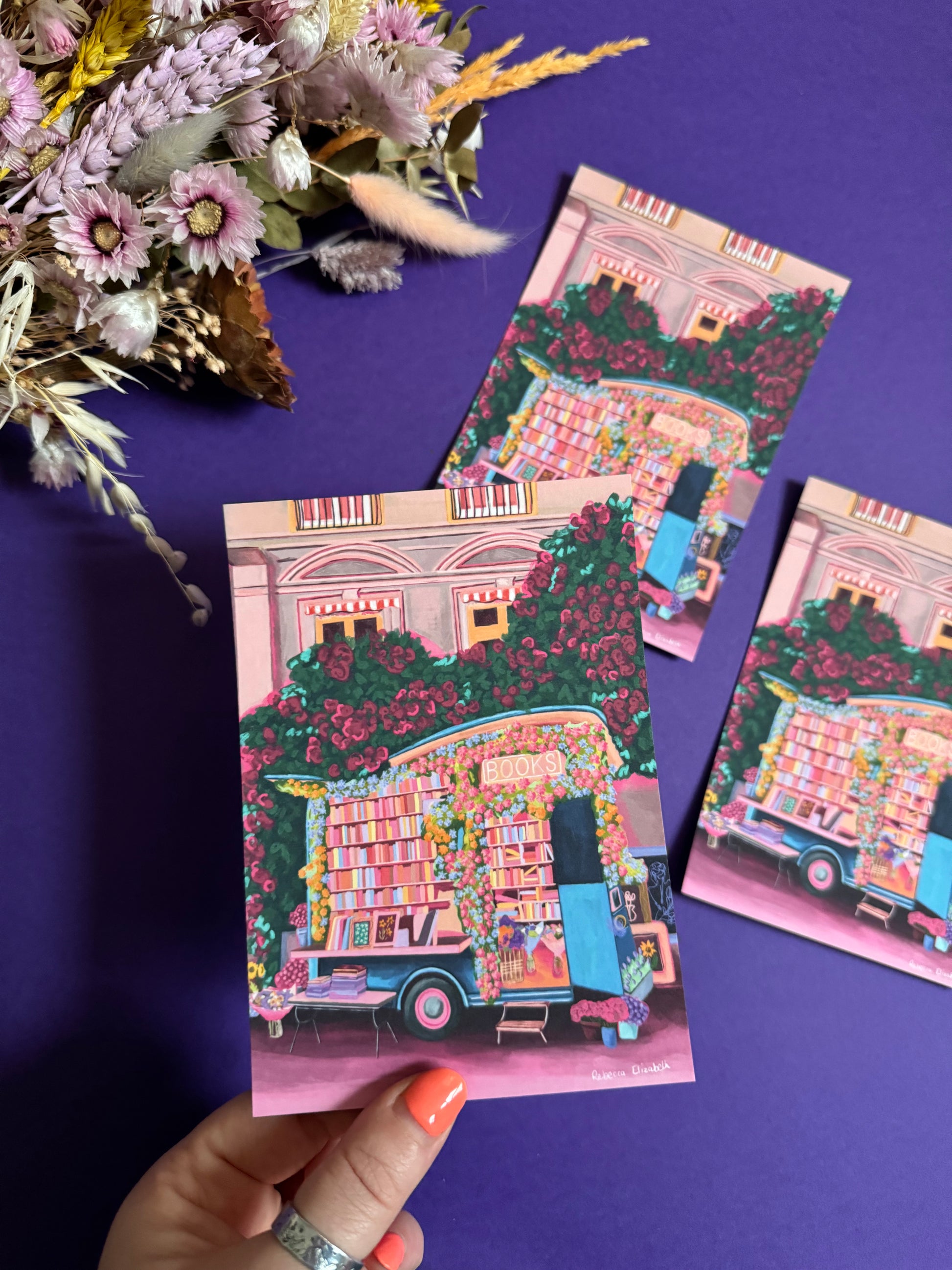 Illustration of a van opened up showing lots of bookshelves stacked with books, art frames out front and lots of blossoming pink, orange and blue flowers over the top. A big tree with pink roses over hangs and behind sits old fashioned buildings with quaint architecture and balconies. A whimsical fantasy print. 