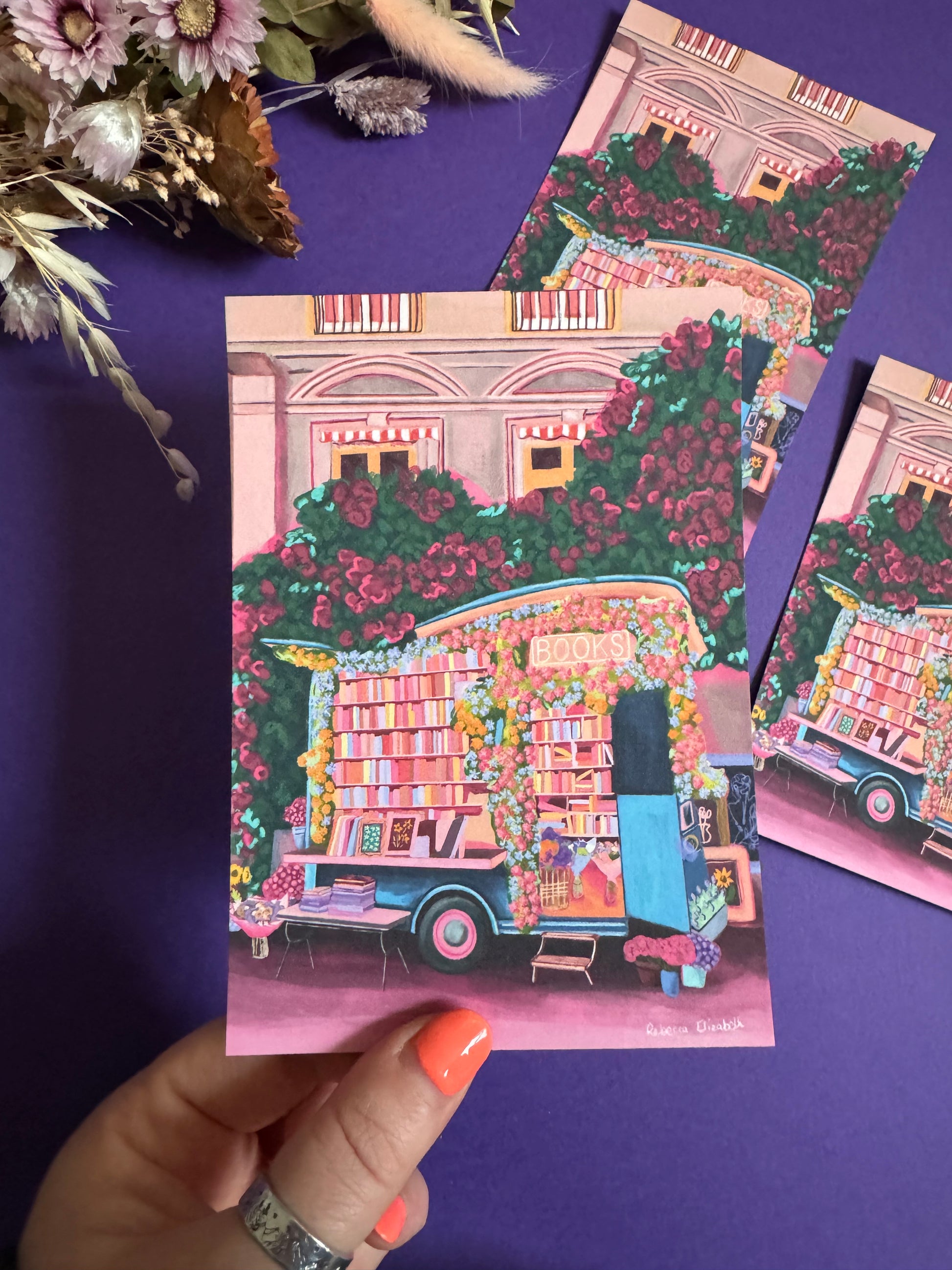 Illustration of a van opened up showing lots of bookshelves stacked with books, art frames out front and lots of blossoming pink, orange and blue flowers over the top. A big tree with pink roses over hangs and behind sits old fashioned buildings with quaint architecture and balconies. A whimsical fantasy print. 