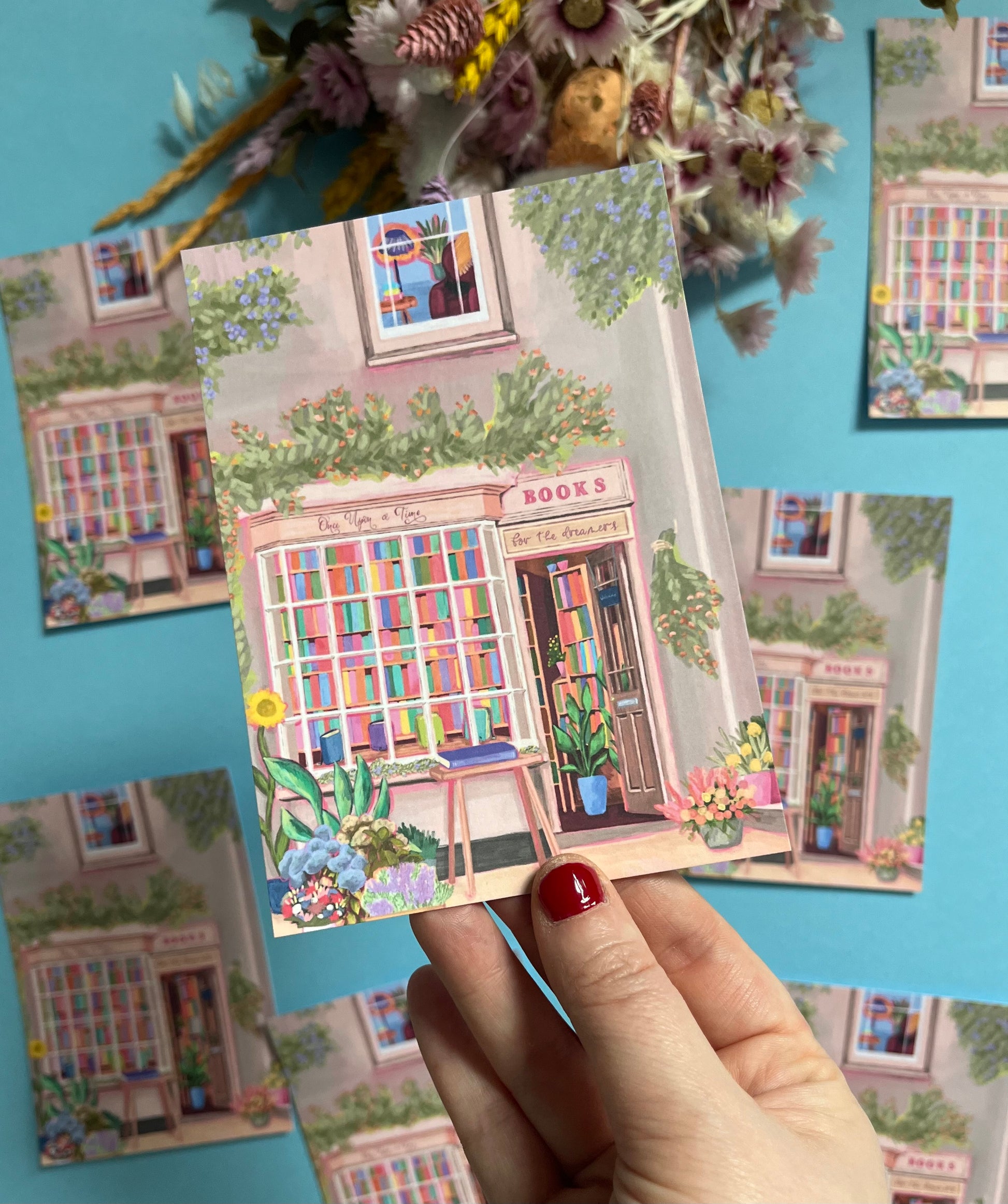 An illustration of a cute bookshop, the window shows the bookshelves inside full of bright coloured book spines. There are books placed stood up at the front. Outside the building is a soft colour with lots of greenery in soft pale greens and orange and lilac flowers. In front of the building there are plant pots and flowers. There is a small signature in white the corner.  Designed by Rebecca Gibbs on a postcard at Rebecca Elizabeth Draws.