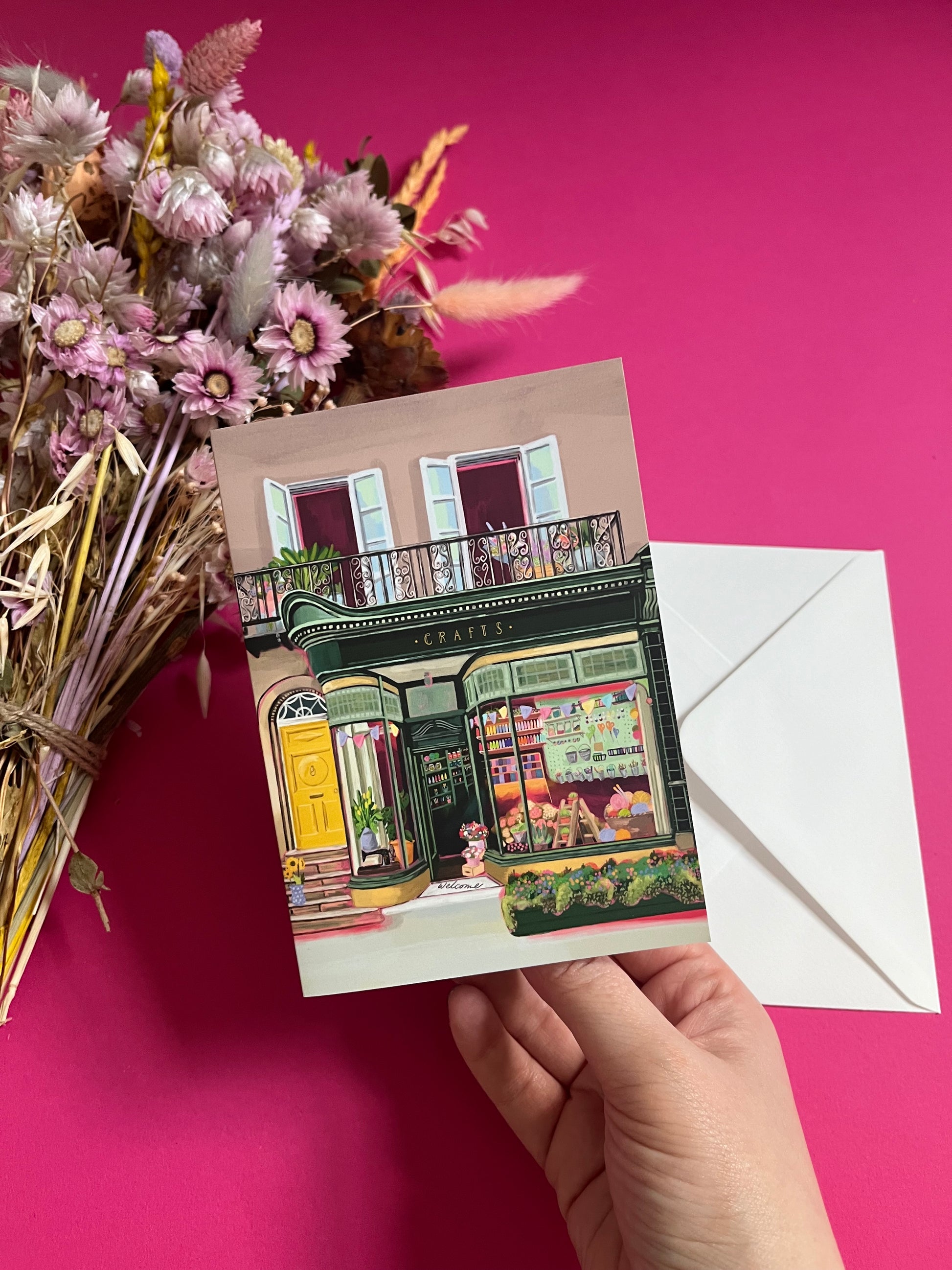 A colourful illustration of a craft shop with the store front in green and looking through the window and at all the goodies. Sewing, drawing, painting, we have it all here at the craft shop. On a greeting card with a green back & logo.