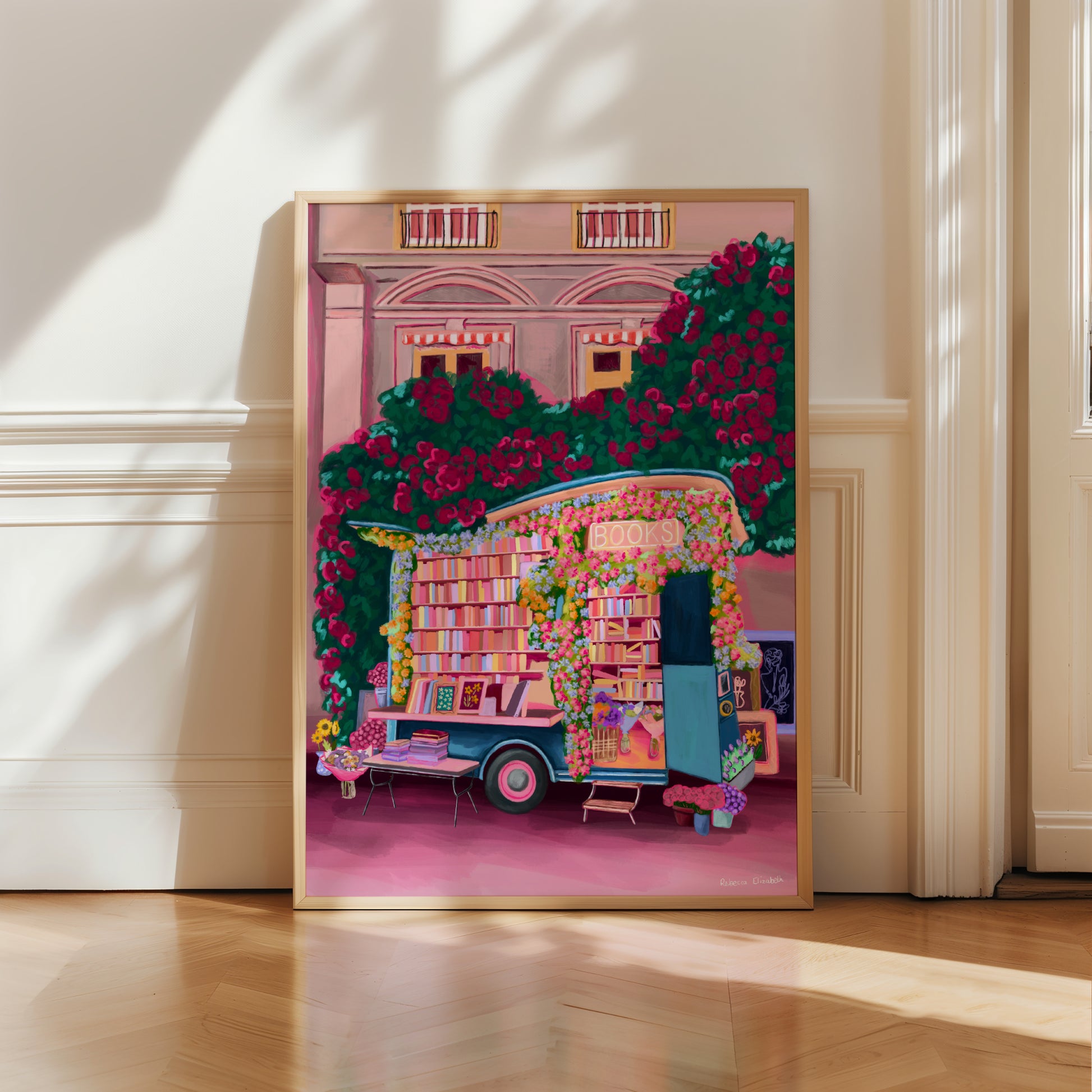 Illustration of a van opened up showing lots of bookshelves stacked with books, art frames out front and lots of blossoming pink, orange and blue flowers over the top. A big tree with pink roses over hangs and behind sits old fashioned buildings with quaint architecture and balconies. A whimsical fantasy print. 