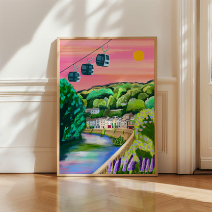 A vibrant and colourful illustration of the beautiful Derbyshire Dales village, Matlock Bath. This illustration features the famous cable cars in the top left hand corner. Then a look across from the river, with the houses and shops set against the big hills. Lots of greenery and a bright pink sky with a bright shining sun. There is a small signature in white the corner.  Designed by Rebecca Gibbs at Rebecca Elizabeth Draws.
