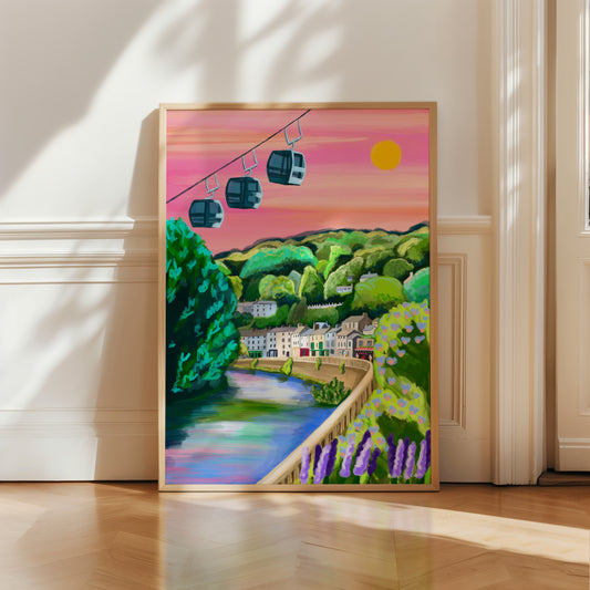 A vibrant and colourful illustration of the beautiful Derbyshire Dales village, Matlock Bath. This illustration features the famous cable cars in the top left hand corner. Then a look across from the river, with the houses and shops set against the big hills. Lots of greenery and a bright pink sky with a bright shining sun. There is a small signature in white the corner.  Designed by Rebecca Gibbs at Rebecca Elizabeth Draws.