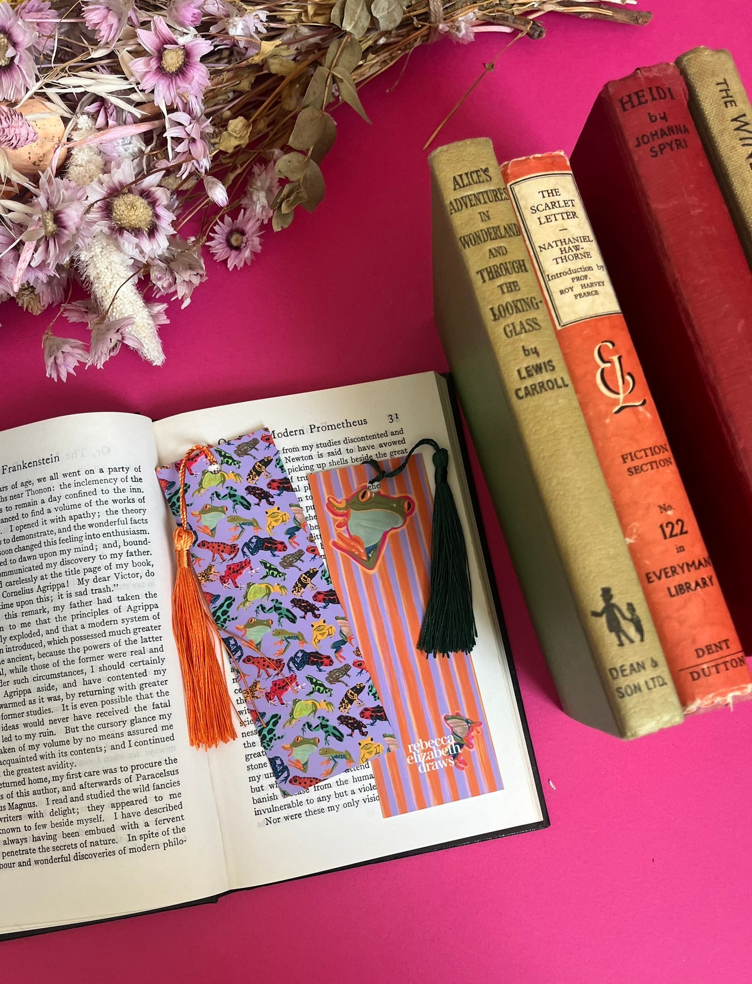 An illustration of frogs on a bookmark. A colourful frog pattern on a purple background on the front and a purple and orange stripe pattern on the back featuring some sneaky peak frogs alongside the Rebecca Elizabeth draws logo. Designed by Rebecca Gibbs at Rebecca Elizabeth Draws.