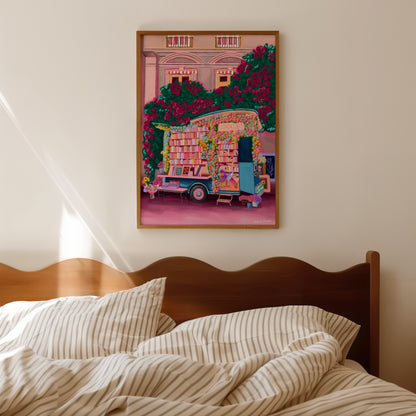 Illustration of a van opened up showing lots of bookshelves stacked with books, art frames out front and lots of blossoming pink, orange and blue flowers over the top. A big tree with pink roses over hangs and behind sits old fashioned buildings with quaint architecture and balconies. A whimsical fantasy print. 
