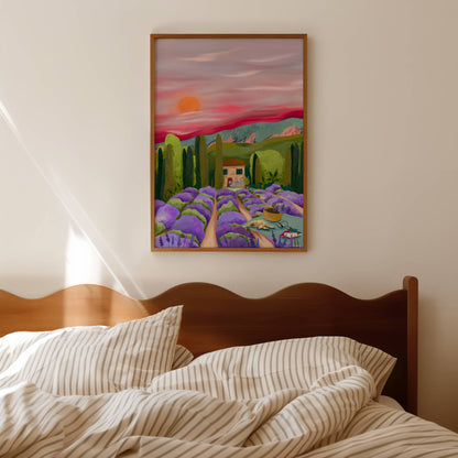 Lavender fields Provence, France. Purple lavender flowers leading to the centre. A picnic blanket with a dog, camera & books alongside a basket full of picked flowers. The centre features a house, big green trees & mountains set against a pink sky.