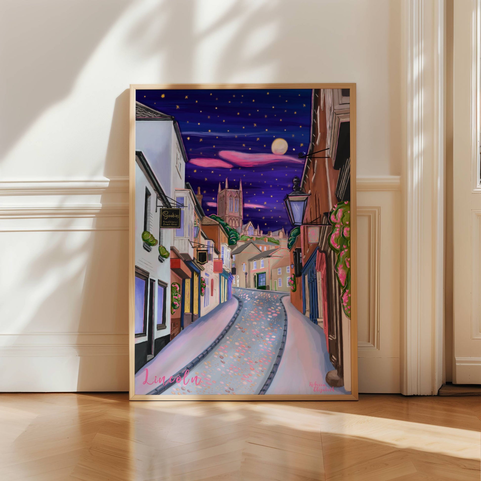 illustration of steep hill in Lincoln, Lincolnshire England UK. IN the background sits Lincoln Cathedral in soft browns & pinks. the sky is a night sky in deep blue with pink cloud tendrils & glittering stars & moon. in the foreground the shops that can be found on steep hill, with a mixture of old fashioned buildings and modern signage. The are pink highlights throughout and little pops of greenery with pink flowers.