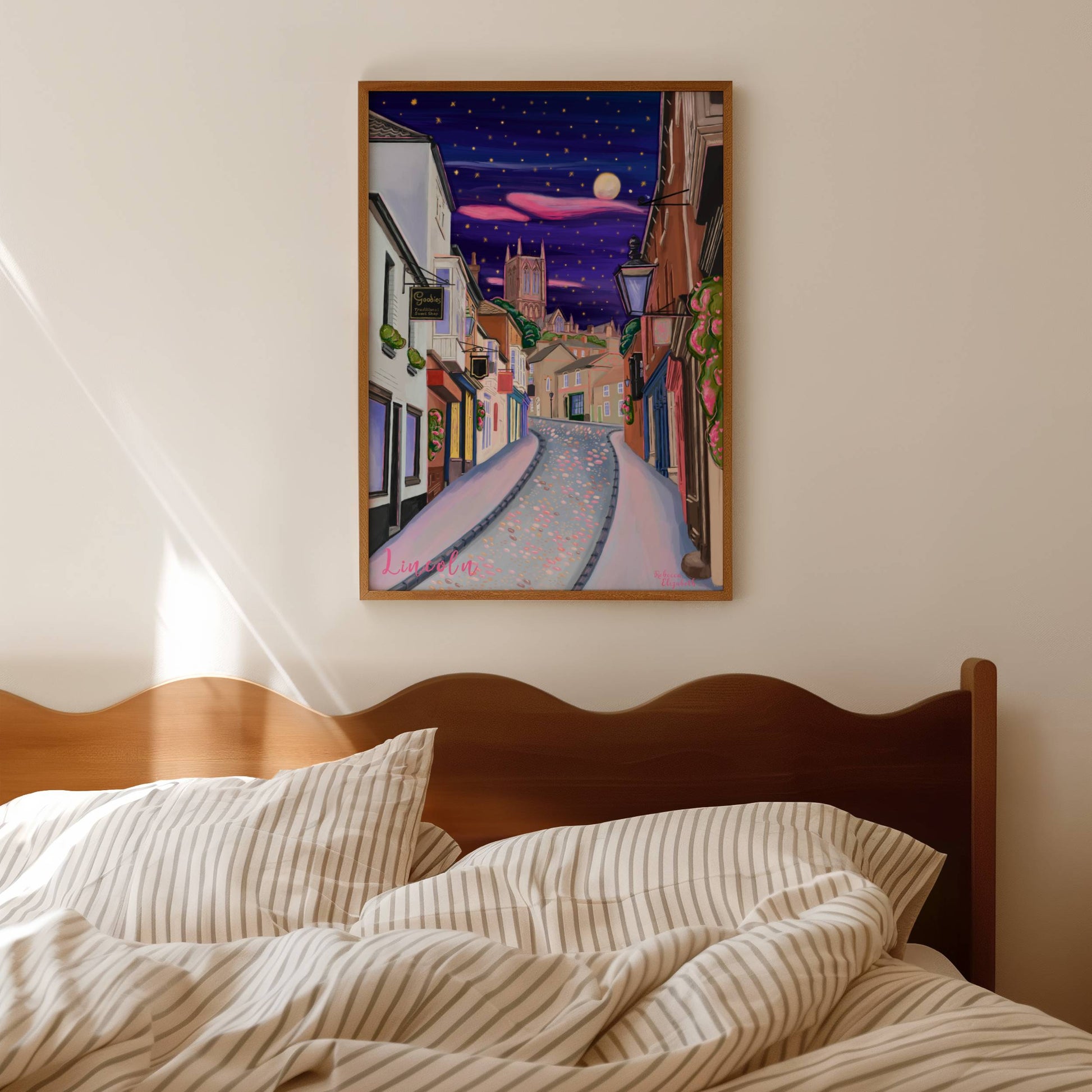 illustration of steep hill in Lincoln, Lincolnshire England UK. IN the background sits Lincoln Cathedral in soft browns & pinks. the sky is a night sky in deep blue with pink cloud tendrils & glittering stars & moon. in the foreground the shops that can be found on steep hill, with a mixture of old fashioned buildings and modern signage. The are pink highlights throughout and little pops of greenery with pink flowers.