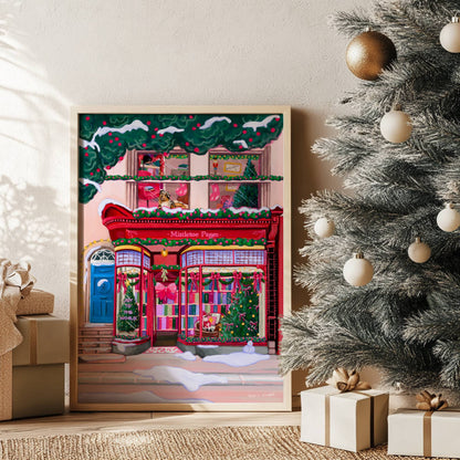 A Christmas bookshop store front illustration in red with soft whimsical colour books, festive decorations & a look upstairs inside the window at a cosy home with a dog. There is snow fallen around & a festive holly & berry tree hanging over the top
