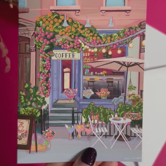 A coffee shop illustrated card with a blue exterior, an outside table and umbrella and building windows surrounding. This illustration is filled with lots of plants and flowers and features cups of coffee and baked goods. Illustrated by Rebecca Elizabeth Draws. 