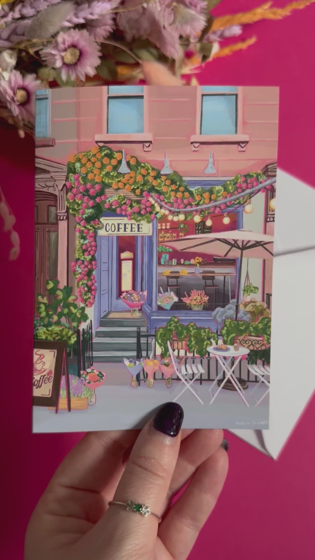 A coffee shop illustrated card with a blue exterior, an outside table and umbrella and building windows surrounding. This illustration is filled with lots of plants and flowers and features cups of coffee and baked goods. Illustrated by Rebecca Elizabeth Draws. 