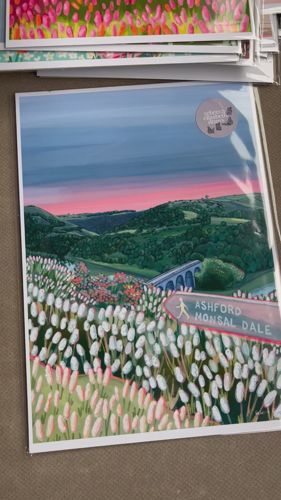 An illustration of Monsal Head in Derbyshire, Peak District. Rolling green hills filled with trees, an old viaduct over the river Wye and white flowers in the foreground. The sky is a setting sun with pink on the horizon fading into dark blue grey. 