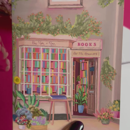 An illustration of a cute whimsical bookshop, the window shows the bookshelves inside full of bright coloured book spines with lots of flowers. The back of the card is pink with the Rebecca Elizabeth Draws logo. 