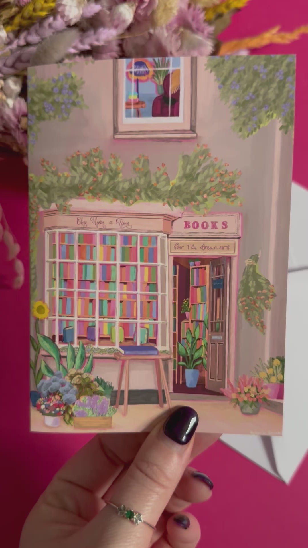 An illustration of a cute whimsical bookshop, the window shows the bookshelves inside full of bright coloured book spines with lots of flowers. The back of the card is pink with the Rebecca Elizabeth Draws logo. 