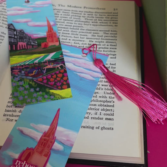 An illustration of chesterfield on a bookmark. A summer illustration featuring the famous crooked spire, market stalls and Tudor buildings. Soft blue sky with fluffy clouds, this art piece is a soft representation of the market town. The back features the same blue sky and clouds and a small crooked spire with the Rebecca Elizabeth draws logo. Designed by Rebecca Gibbs at Rebecca Elizabeth Draws.