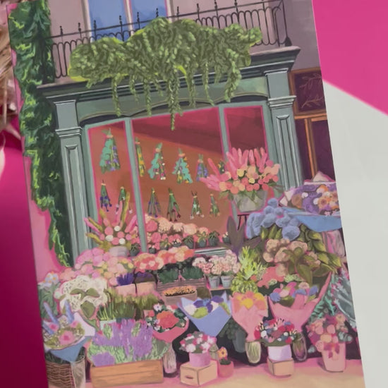 Greeting card of a beautiful flower shop, the window shows the flowers drying inside and outside there are rows of bunches of flowers and pots of plants. Soft brown and pink with bright highlights throughout. With a pink back piece & logo.