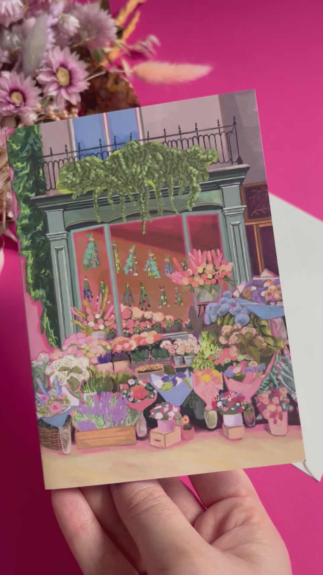 Greeting card of a beautiful flower shop, the window shows the flowers drying inside and outside there are rows of bunches of flowers and pots of plants. Soft brown and pink with bright highlights throughout. With a pink back piece & logo.