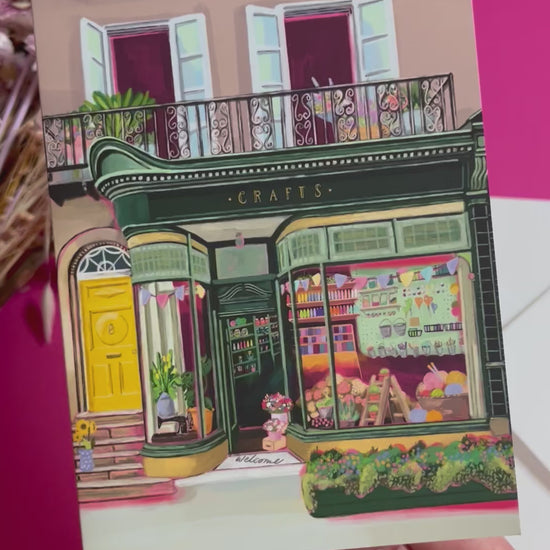 A colourful illustration of a craft shop with the store front in green and looking through the window and at all the goodies. Sewing, drawing, painting, we have it all here at the craft shop. On a greeting card with a green back & logo.