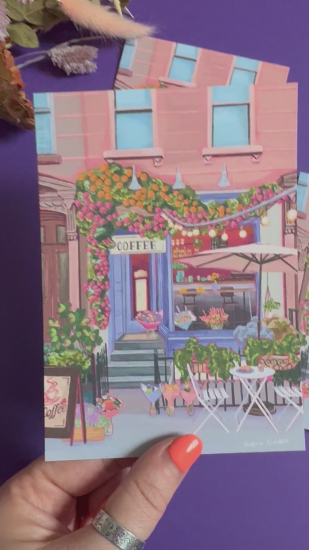 A coffee shop illustration with a blue exterior, an outside table and umbrella and building windows surrounding. This illustration is filled with lots of plants and flowers and features cups of coffee and baked goods. Illustrated by Rebecca Elizabeth Draws. 