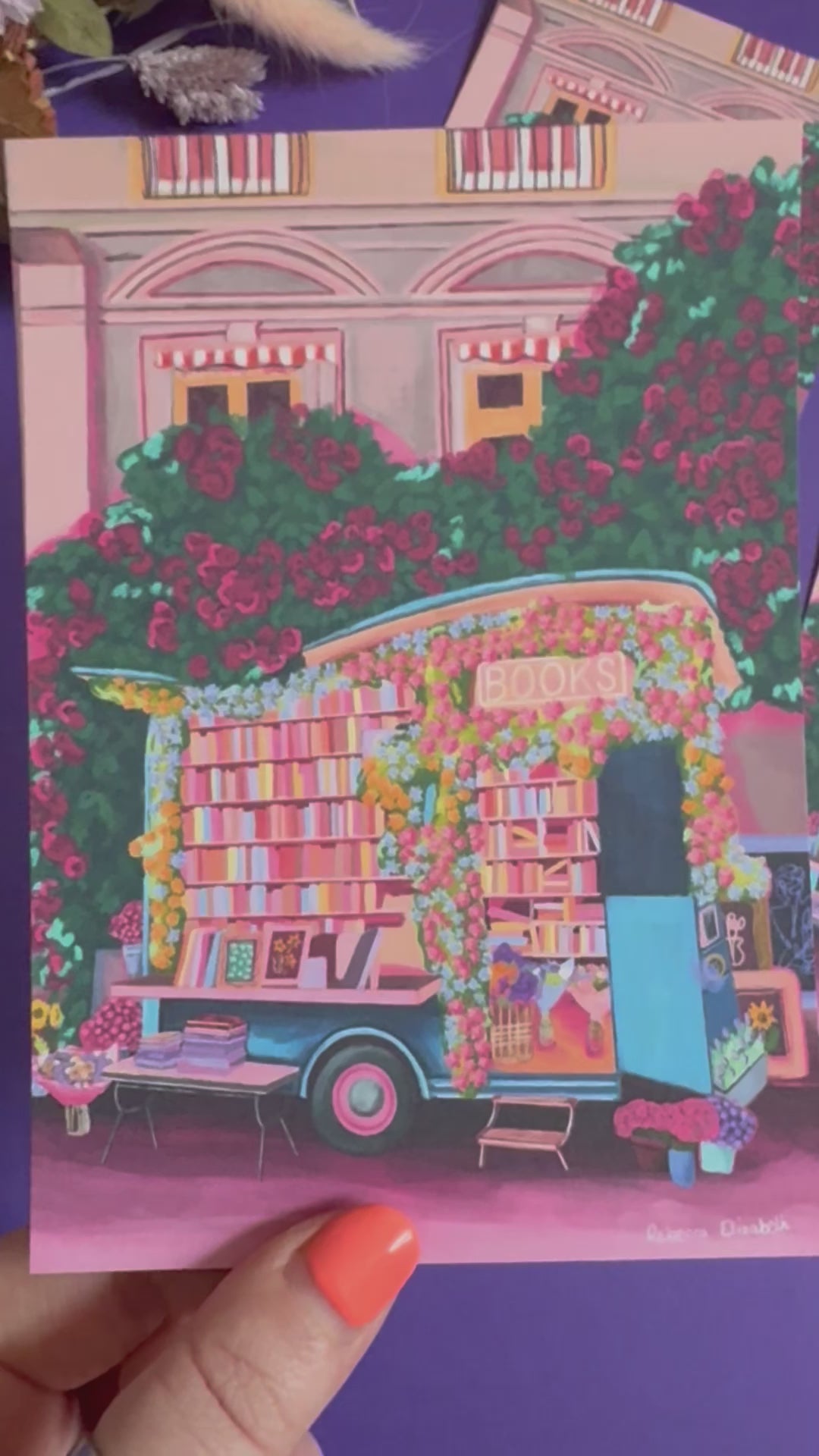 Illustration of a van opened up showing lots of bookshelves stacked with books, art frames out front and lots of blossoming pink, orange and blue flowers over the top. A big tree with pink roses over hangs and behind sits old fashioned buildings with quaint architecture and balconies. A whimsical fantasy print. 