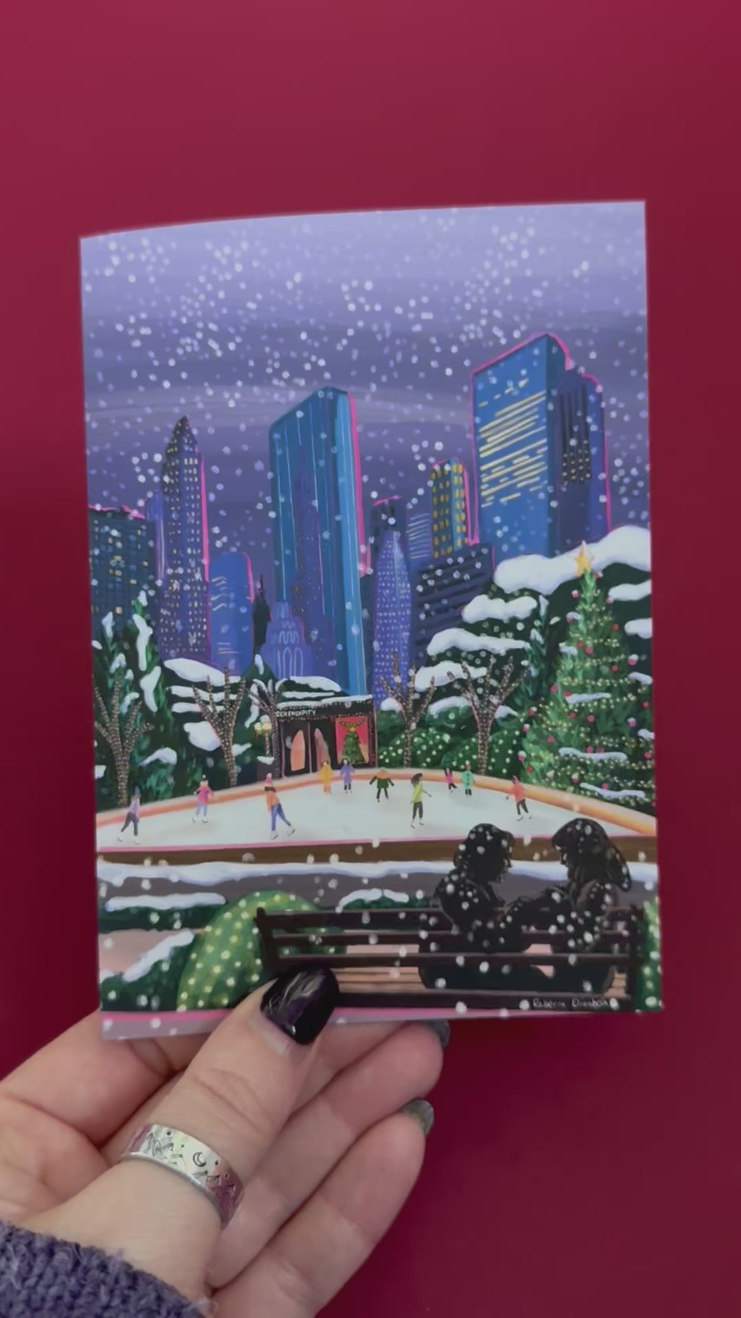 Christmas Illustration inspired by the film Serendipity. The main stars are silhouetted at the front of this illustration with the new York skyline behind them, at the base of the high rise buildings are Christmas trees, the serendipity shop and an ice skate rink with people on. A christmas card with a blue back designed by Rebecca Gibbs at Rebecca Elizabeth Draws.