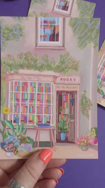 An illustration of a cute bookshop, the window shows the bookshelves inside full of bright coloured book spines. There are books placed stood up at the front. Outside the building is a soft colour with lots of greenery in soft pale greens and orange and lilac flowers. In front of the building there are plant pots and flowers. There is a small signature in white the corner.  Designed by Rebecca Gibbs on a postcard at Rebecca Elizabeth Draws.