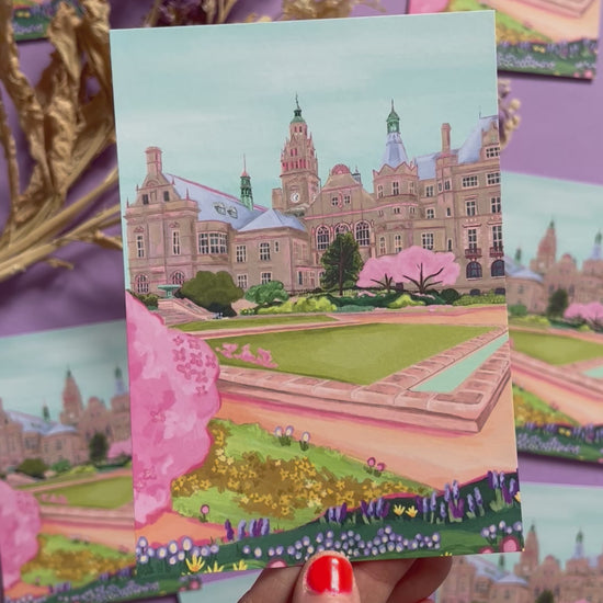 An illustration of the view of the Peace Gardens, Sheffield city centre in springtime. A soft brown building of Sheffield’s town hall with trees in front and pink blossom trees in soft pinks. The foreground features the peace garden with patches of grass and then flowers in yellows and purples underneath another pink blossom tree in the front and a baby blue sky. There is a small signature in white the corner.  Designed by Rebecca Gibbs on a postcard at Rebecca Elizabeth Draws.