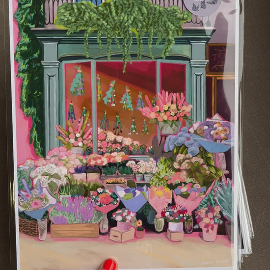 An illustration of a cute bookshop, the window shows the bookshelves inside full of bright coloured book spines. There are books placed stood up at the front. Outside the building is a soft colour with lots of greenery in soft pale greens and orange and lilac flowers. In front of the building there are plant pots and flowers. There is a small signature in white the corner.  Designed by Rebecca Gibbs at Rebecca Elizabeth Draws.