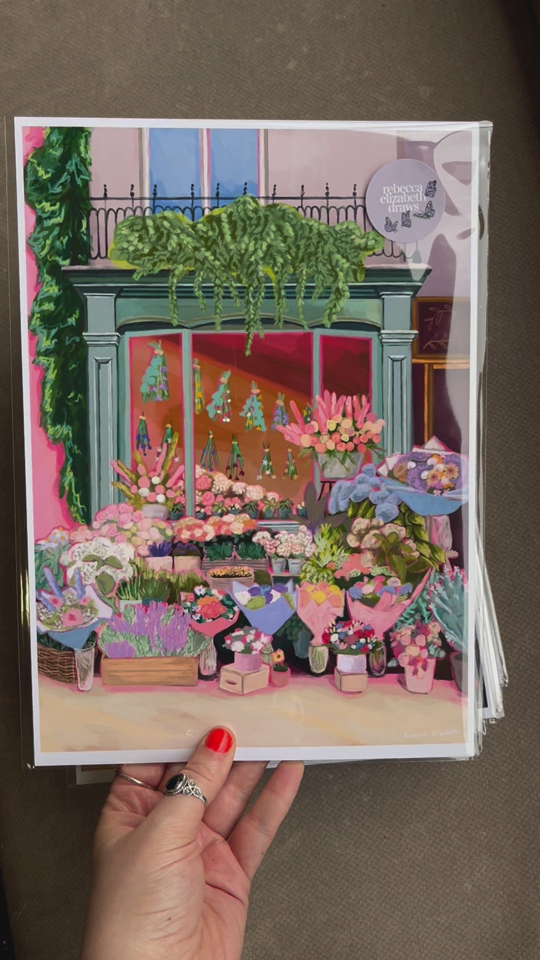 An illustration of a cute bookshop, the window shows the bookshelves inside full of bright coloured book spines. There are books placed stood up at the front. Outside the building is a soft colour with lots of greenery in soft pale greens and orange and lilac flowers. In front of the building there are plant pots and flowers. There is a small signature in white the corner.  Designed by Rebecca Gibbs at Rebecca Elizabeth Draws.