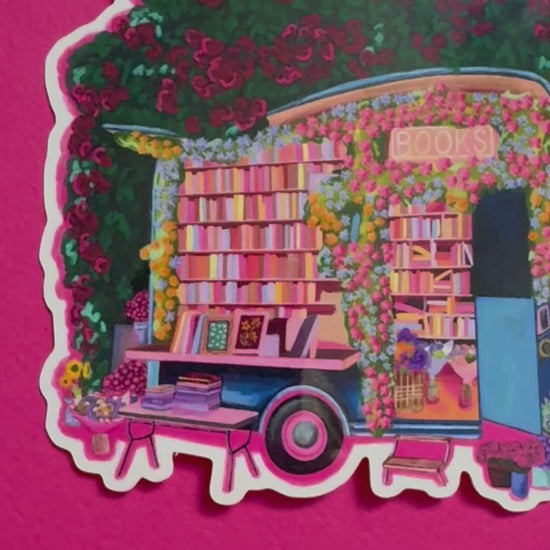 Illustration of a van opened up showing lots of bookshelves stacked with books, art frames out front and lots of blossoming pink, orange and blue flowers over the top. A big tree with pink roses over hangs and behind sits old fashioned buildings with quaint architecture and balconies. A whimsical fantasy sticker.