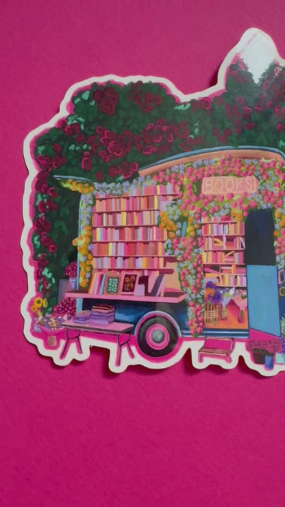 Illustration of a van opened up showing lots of bookshelves stacked with books, art frames out front and lots of blossoming pink, orange and blue flowers over the top. A big tree with pink roses over hangs and behind sits old fashioned buildings with quaint architecture and balconies. A whimsical fantasy sticker.