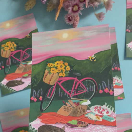 An illustration of a pink bicycle resting up by a picnic blanket filled with wine, loaves of bread, strawberries, picked sunflowers and a book. There is a cat sleeping and behind is pink and purple flowers against the greenery. There is a bumble bee and the sky is pink and grey with a small yellow sun. There is a small signature in white the corner.  Designed by Rebecca Gibbs on a postcard at Rebecca Elizabeth Draws.