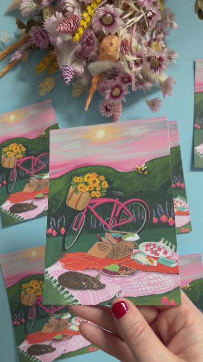 An illustration of a pink bicycle resting up by a picnic blanket filled with wine, loaves of bread, strawberries, picked sunflowers and a book. There is a cat sleeping and behind is pink and purple flowers against the greenery. There is a bumble bee and the sky is pink and grey with a small yellow sun. There is a small signature in white the corner.  Designed by Rebecca Gibbs on a postcard at Rebecca Elizabeth Draws.