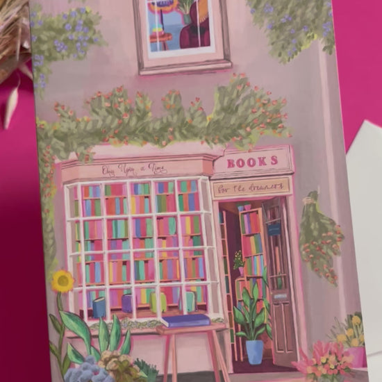 An illustration of a cute whimsical bookshop, the window shows the bookshelves inside full of bright coloured book spines with lots of flowers. The back of the card is pink with the Rebecca Elizabeth Draws logo. 