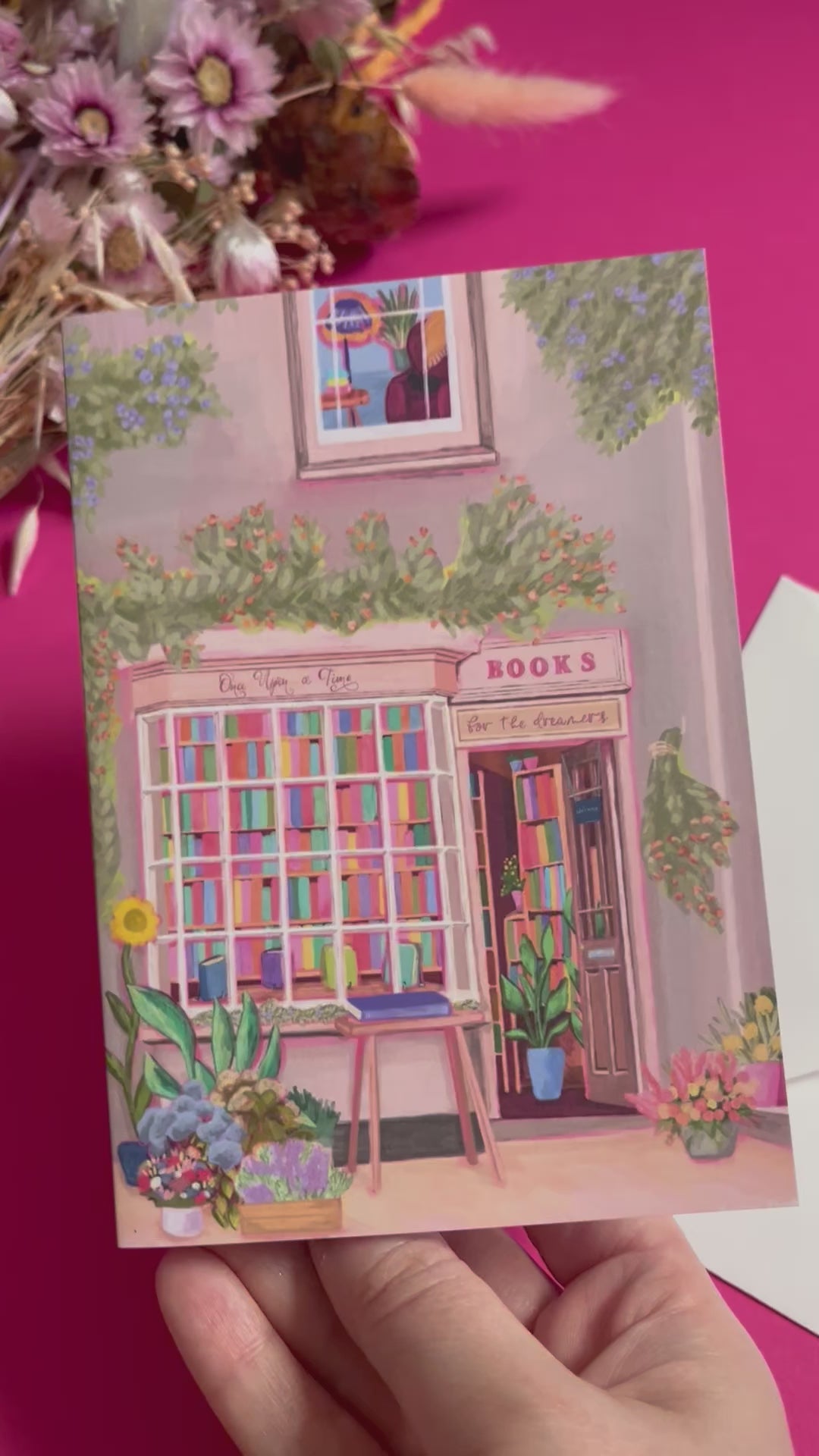 An illustration of a cute whimsical bookshop, the window shows the bookshelves inside full of bright coloured book spines with lots of flowers. The back of the card is pink with the Rebecca Elizabeth Draws logo. 