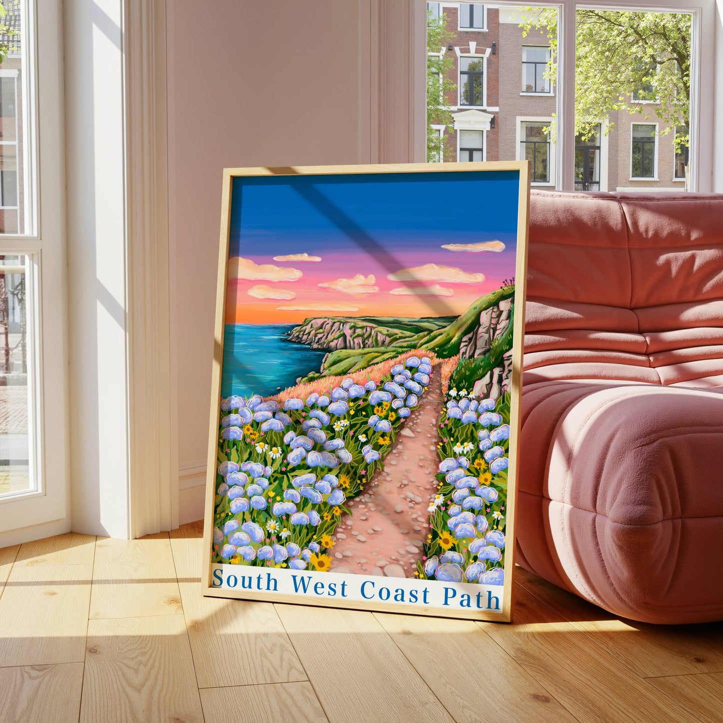 Framed artwork of a scenic path with flowers in a room with large windows and a red sofa.